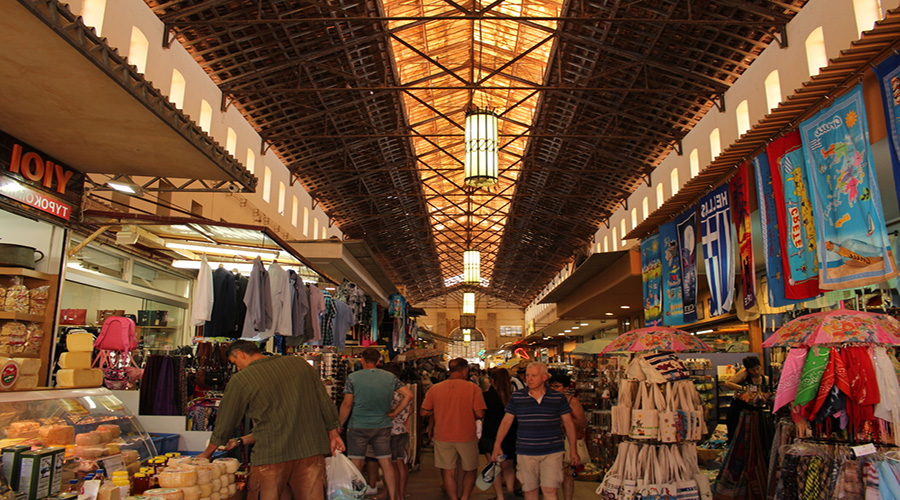 Interior del mercado municipal cubierto de Chania, con puestos de quesos, ropa y recuerdos bajo una estructura de hierro y paseantes en el pasillo central